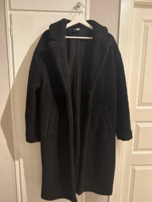 Svart lång teddykappa från H&M - Mysig svart teddykappa från H&M i lång modell med krage och knappar framtill. Jackan har två sidofickor och är tillverkad i fluffigt teddy-material som håller dig varm. Perfekt för dig som gillar oversized och chill vibe.