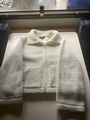 Vit teddyjacka från 157, XS - Mysig vit teddyjacka från 157 i storlek XS. Jackan har två stora fickor framtill, dragkedja och en klassisk krage. Materialet är fluffigt och ger en cool, avslappnad vibe. Perfekt för dig som gillar stilren och enkel design.