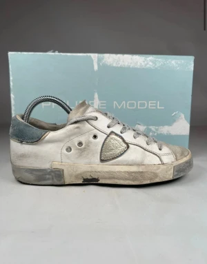 Philippe Model vita sneakers i skinn - Snygga vita sneakers från Philippe Model med grå detaljer och silverfärgad skinnpatch på sidan. Skorna har klassisk låg profil, rund tå och snörning. Ovandelen är i skinn och mocka, med slitna detaljer för en cool, urban look. Perfekta för dig som gillar streetstyle. Skriv om ni har frågor eller funderingar!😊