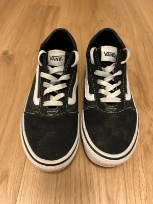 Svarta Vans  - Klassiska Vans Old Skool sneakers i svart mocka och canvas med vit sidorand och vita snören. Skorna har en platt gummisula med det ikoniska rutmönstret undertill och rund tå. Perfekta för dig som gillar streetstyle och skatekänsla.