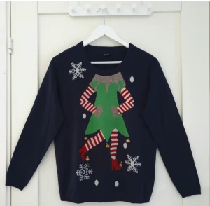 Mörkblå jultröja med elf-motiv - Rolig mörkblå jultröja från New Look med ett stort elf-motiv framtill, glittriga detaljer och små bjällror. Tröjan har långa ärmar och snöflingor som dekoration. Perfekt för julmys och temadagar!