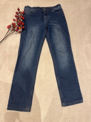 Blå jeans från Lyle & Scott, 14-15 år - Snygga blå jeans från Lyle & Scott med klassisk femficksdesign och gul broderad logga på bakfickan. Jeansen har raka ben och normal passform, med bruna detaljer vid midjan. Perfekt för dig som gillar stilrena och tidlösa jeans. Storlek 164