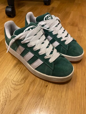 Adidas Campus gröna sneakers 42 2/3 - Gröna Adidas Campus sneakers i mocka med vita detaljer och klassiska tre ränder på sidan. Skorna har vit sula, breda vita skosnören och Adidas logga på plösen. Perfekt för dig som gillar retrostil och vill sticka ut med färg.