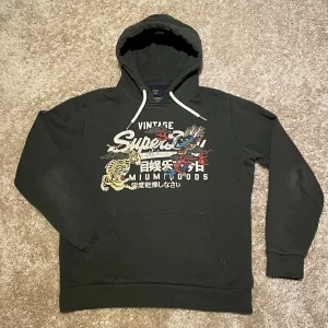 Superdry hoodie - En riktigt fet Superdry hoodie med ett unikt och snyggt tryck med japanska detaljer, den är mörkgrön/olivgrön med små detaljer som en liten patch på armen och liten text på snörerna. Den har inga defekter eller någon sorts skada. Kom pm för mått, inga skambud 