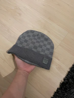 Grå Iv mössa - Snygg grå mössa från Iv med diskret rutmönster och ribbad kant. Märkesplakett i grått framtill. Stickad i mjukt material som håller dig varm och ger en lyxig vibe. Perfekt för dig som gillar designeraccessoarer.