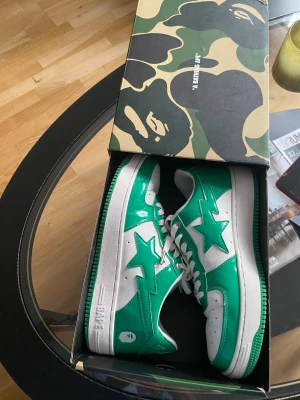 BAPE STA vita sneakers med grön design  - Säljer ett par BAPE APE sneakers i grön och vit färg med klassisk stjärndesign på sidan. Skorna har blank finish, vita snören och perforerad tåbox. Ikonisk BAPE-logga på hälen och metall-detalj på snörningen. Kommer i originalkartong med camomönster. Knapptanvända med lite slitage som synlig på bilderna . Säljes helt rena och fräscha utan konstiga lukt , sulan också rent. Inga hål . Normal storlek 43 ( 10 enligt lapp) 