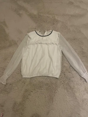 Vit blus med spets och transparenta ärmar - Säljer en vit blus med transparenta långärmar och elastisk nederkant. Blusen har spetsdetaljer vid axlar och bröst, samt en svart kant vid halsen. Perfekt för dig som gillar romantisk och söt stil med extra detaljer.