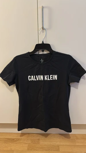 Svart Calvin Klein t-shirt S - Svart t-shirt från Calvin Klein med vit logotyp tryckt framtill och CK-detalj i nacken. Modellen är klassisk med rund hals och korta ärmar. Materialet är mjuk bomull med lite stretch för skön passform. Perfekt för dig som gillar stilrena och ikoniska plagg.