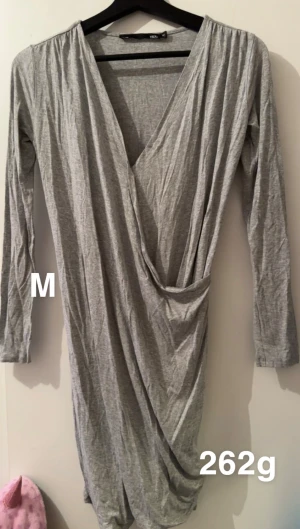 Grå omlottklänning med v-ringning - Snygg grå omlottklänning från lager 157 i storlek M. Klänningen har lång ärm, djup v-ringning och draperad front som ger en cool och avslappnad look. Perfekt för dig som gillar stilrena och enkla plagg.🌸 Fint använt skick. Storlek m