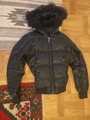 Svart 2000s  pufferjacka med fuskpäls från Bomboogie - Svart pufferjacka från Bomboogie med huva och fuskpäls. Jackan har ribbade muddar, dragkedja framtill och två fickor med tryckknappar. Perfekt för kalla dagar och har en snygg, sportig look. Köpt på secondhand i Grekland 2011. Äkta vintage och 2000s stil