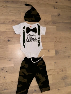 Camouflage babybyxor med snörning - Säljer ett par coola babybyxor i camouflage-mönster med vit snörning i midjan. Byxorna har en loose passform och är perfekta för små äventyrare. Materialet är mjukt och bekvämt, troligen bomull eller polyester. Matcha med bodyn för en komplett look!Ett set med en vit babybody med tryck "Ladies, I have arrived", ett par camouflage byxor och en matchande mössa. 🎁 