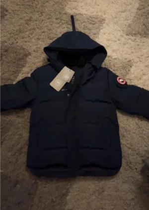 Mörkblå dunjacka från Canada Goose - Säljer en mörkblå dunjacka från Canada Goose med huva och klassisk logga på ärmen. Jackan har quiltad design och dragkedja framtill. Perfekt för kalla dagar och har en clean look som funkar till det mesta.