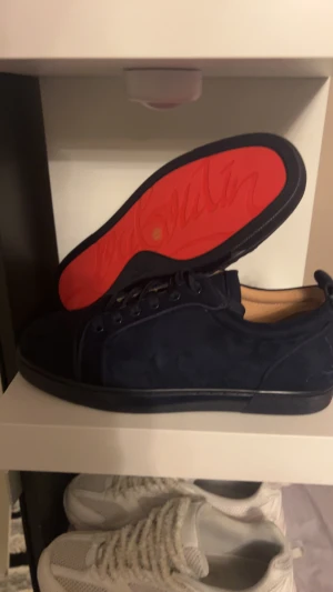 Mörkblå mocka sneakers från Louboutin - Säljer ett par stilrena mörkblå sneakers från Christian Louboutin i mjuk mocka. Skorna har den ikoniska röda sulan med Louboutin-logga, rund tå och snörning. Perfekt för dig som vill ha en lyxig och unik touch på din outfit.