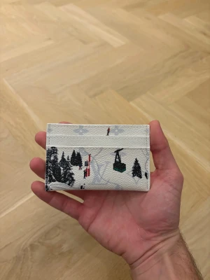 Vit plånbok med vintermotiv - Vit korthållare i läder med tryckt vinterlandskap, skidåkare, lift och granar. Smidig och stilren accessoar med flera kortfack, perfekt för dig som gillar unika detaljer och vintriga vibes.