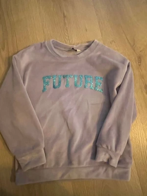 Ljuslila sweatshirt med FUTURE-tryck - Ljuslila sweatshirt i mjukt material med glittrigt turkost FUTURE-tryck på bröstet. Tröjan har rund halsringning och muddar vid ärmslut och nederkant. Perfekt för dig som gillar statement-plagg och vill sticka ut lite extra.