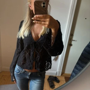 Svart blus med snörning och hålmönster - Svart blus med v-ringning och snygg snörning framtill. Blusen har långa, vida ärmar och är gjord i ett genombrutet bomullstyg med broderade detaljer. Perfekt för dig som gillar boho-vibe och vill sticka ut lite extra.