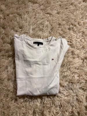 Vit t-shirt från Tommy Hilfiger - Vit t-shirt från Tommy Hilfiger med klassisk rund hals och korta ärmar. T-shirten har en liten broderad logga på bröstet i rött, vitt och blått. Tillverkad i mjuk bomull för en skön känsla. Enkel och stilren design som funkar till allt.