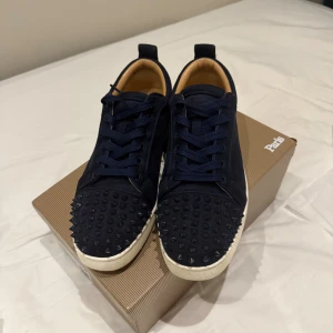 Mörkblå Christian Louboutin sneakers - Snygga mörkblå sneakers från Christian Louboutin med ikoniska röda sulor och svarta nitar på tån. Skorna har snörning, rund tå och vit platt sula. Tillverkade i mocka med lyxig känsla och cool attityd. Perfekt för dig som vill sticka ut med exklusiva detaljer. 3 spikar av