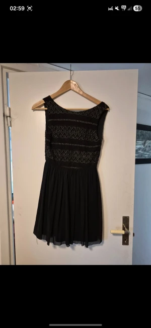 Svart kort klänning med broderi - Supersnygg svart kort klänning med guldigt broderi på överdelen. Klänningen har ärmlös design, markerad midja och lätt utställd kjol i tunt tyg. Djup rygg med knytband ger en extra detalj. Perfekt för dig som gillar unika detaljer och stilren look.