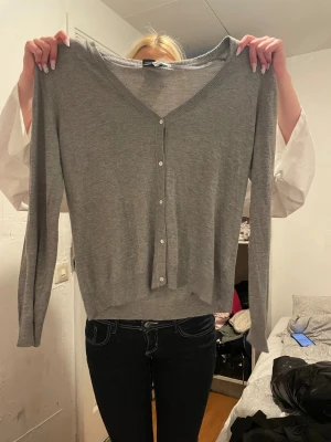 Grå kofta från KappAhl, stl 44/46 - Snygg grå kofta från KappAhl i storlek 44/46. Den har v-ringning, långa ärmar och knäppning framtill med små knappar. Perfekt att slänga över en t-shirt eller linne. Mjuk och skön, passar till allt.