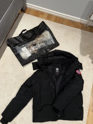 Svart pufferjacka från Canada Goose - Svart pufferjacka från Canada Goose i storlek M. Jackan har en stor huva, klassisk logga på ärmen och dragkedja framtill. Fodret är dun och yttertyget är slitstarkt syntetmaterial. Perfekt för kalla vinterdagar och har en clean, modern look.