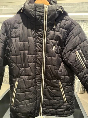 Svart skid-& vinterjacka från Peak  - Svart skid-& vinterjacka från Peak Performance med quiltat mönster, hög krage och huva med dragsko. Jackan har vita dragkedjor, flera fickor med dragkedja, snölås mm. Perfekt för kalla dagar och riktigt snygg. 
