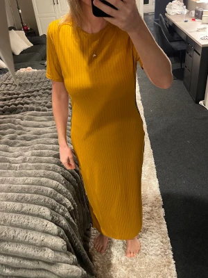 Ribbad gul långklänning med textband - Säljer en cool ribbad långklänning i starkt gul färg med korta ärmar och rund hals. Klänningen har ett svart textband med vit text längs sidan som ger en sportig vibe. Materialet är mjukt och stretchigt, perfekt för dig som gillar att sticka ut.