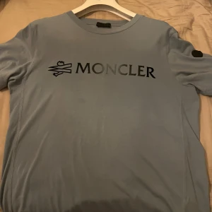  Moncler t-shirt - Snygg ljusblå t-shirt från Moncler med svart logotyp på bröstet och en svart patch på ärmen. Rund hals och korta ärmar. Använd en gång, så i perfekt skick. Köpt på moncler store i Stockholm. Tags, kvitton och dustbag medföljer. Nypris 3090kr.