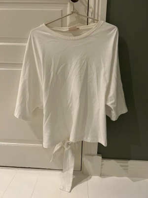 Vit topp med knyt från Nelly - Vit topp från Nelly i mjuk bomull med rund halsringning och trekvartsärmar. Den har även knyt därbak som man kan knyta själv. Skulle säga att den passar de flesta eftersom att man kan nästa bestämma hur hårt knytet man vill ha den. Aldrig använd