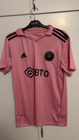 Inter Miami Messi tröja Adidas L - Säljer en rosa Inter Miami t-shirt från Adidas med Messi 10-tryck på ryggen. Tröjan har svarta detaljer, klubbmärke på bröstet och klassiska Adidas-ränder på axlarna. Slim fit-modell i lätt polyester, perfekt för fotboll eller att bära casual.