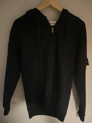 Stone Island hoodie - Svart hoodie från Stone Island med klassisk logotyp på ärmen. Tröjan har huva, dragkedja framtill och två fickor. Materialet är mjuk bomull och passformen är normal. Perfekt för dig som gillar streetwear och vill ha en clean look. Fråga för fler bilder. Obs pris kan diskuteras.