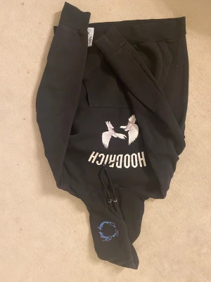 Svart hoodie med vita fåglar från Hoodrich - Svart hoodie från Hoodrich med tryck av två vita fåglar på bröstet och text i blått och vitt. Huva med snörning och blå textdetalj. Långärmad och med en stor ficka framtill, snygga detaljer på hela tröjan. Ganska stor i storleken.