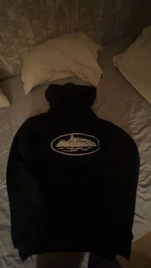 Svart corteiz hoodie  - Svart hoodie från Skeppsholmen med ett stort vitt tryck av ett skepp på bröstet. Tröjan har huva och är långärmad, tillverkad i mjuk bomullsmix som är skön att bära. Perfekt för dig som gillar streetwear och vill ha en unik look.