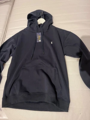 Mörkblå hoodie från Polo Ralph Lauren - Snygg mörkblå hoodie från Polo Ralph Lauren med klassisk logga broderad på bröstet. Tröjan har huva med snörning, stor magficka och ribbade muddar. Materialet är mjuk bomull, perfekt för chill dagar. Passar dig som gillar stilren och enkel design. Tröjan är lite mindre i storleken än L passar mer på en M