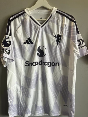 Manchester United bortastäl Cunha #10 - Snygg vit och ljuslila fotbollströja från Adidas med Cunha och nummer 10 på ryggen. Tröjan har korta ärmar, v-ringad krage och coolt geometriskt mönster. Premier League-logga på ärmen och Snapdragon-tryck på bröstet. Perfekt för dig som älskar fotboll och stil.       Är du intresserad är det bara att höra av sig 