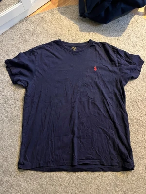 Marinblå t-shirt från Polo Ralph Lauren - Klassisk marinblå t-shirt från Polo Ralph Lauren i bomull. T-shirten har korta ärmar, rund halsringning och den ikoniska röda broderade loggan på bröstet. Perfekt basic-plagg med snygg passform och stilren look.