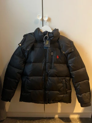 Svart pufferjacka från Polo Ralph Lauren - Säljer en svart pufferjacka från Polo Ralph sopad med röd broderad logga på bröstet. Jackan har SNYGG hög krage, dragkedja framtill, fickor och avtagbar huva. Perfekt för kalla dagar och riktigt clean look.