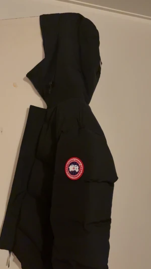 Svart pufferjacka från Canada Goose - Svart pufferjacka från Canada Goose med ikonisk röd och vit logga på ärmen. Jackan har huva med snörning, ribbade muddar och dragkedja framtill. Materialet är slitstark polyester och dunfyllning som håller dig varm under kalla dagar. Perfekt för dig som vill ha en clean och stilren vinterlook.