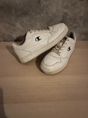 Vita sneakers från Champion Storlek 41 - Snygga vita sneakers från Champion med klassisk låg profil och rund tå. Skorna har snörning och är tillverkade i syntetmaterial med perforerade detaljer på sidorna. Champion-loggan i svart syns tydligt på utsidan. Perfekta för dig som gillar clean och sportig stil.