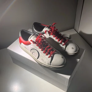 Oliver Cabell vita sneakers med röd detalj - Snygga vita sneakers från Oliver Cabell med röda skosnören och röd häl, samt svart detalj på bakkappan. Skorna har rund tå, platt sula och är tillverkade i skinn. En cirkelformad detalj på sidan ger dem en unik look.