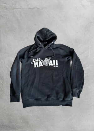 Svart hoodie Cold Hawaii West Wind - Svart hoodie från Lagos med trycket 'Cold Hawaii West Wind' i vitt på bröstet och en grafisk hand. Tröjan har huva med snörning och ribbade muddar vid ärmslut och nederkant. Perfekt för chill dagar och streetstyle.