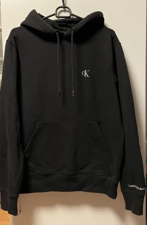 Svart hoodie från Calvin Klein Jeans - Svart hoodie från Calvin Klein Jeans med broderad cK-logga på bröstet och Calvin Klein Jeans-tryck på ärmen. Tröjan har huva med snörning, stor magficka och ribbade muddar. 