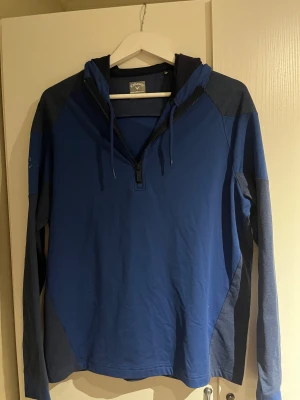 Sällsynt Blå hoodie från Callaway Weather Series - Snygg blå hoodie från Callaway Weather Series med mörkblå detaljer och broderad logga på ärmen. Tröjan har huva med snörning, half zip framtill och långa ärmar. Perfekt för dig som gillar sportig stil och vill ha något bekvämt och funktionellt. Funkar perfekt på höstdagar men även sen sommarkvällar. Skick 10/10 aldrig använd. 