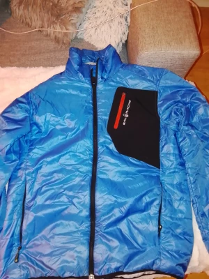 Blå pufferjacka från Sail Racing - Blå pufferjacka från Sail Racing med svart bröstfickaJackan har hög krage, hel dragkedja framtill och två sidofickor med dragkedja. Materialet är nylon och polyester, vilket gör den lätt och skön att bära. Perfekt för kalla dagar.