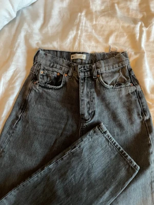 Grå jeans - Säljer ett par grå jeans från Gina Tricot med hög midja och raka ben. Klassisk femficksmodell med silverfärgade nitar och knapp. Jeansen har en lite sliten look och är tillverkade i jeansmaterial. 