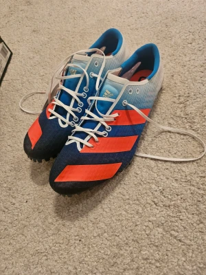 Spikskor adidas  - Adidas Adizero Finesse löparskor med snörning, designade för friidrott. Skorna har en snygg gradient från blått till vitt med röda detaljer och vita skosnören. Ovandelen är i syntetmaterial och medföljer lösa spikar och nyckel för montering. Obs! Något små i storleken.