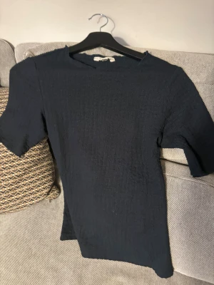 Mörkblå T-shirt  med struktur - Säljer en stilren mörkblå T-shirt  med korta ärmar och lätt strukturerad yta. Toppen har en rak passform och enkel design som gör den lätt att matcha med olika outfits. Perfekt för dig som gillar minimalistisk stil och mörka färger.