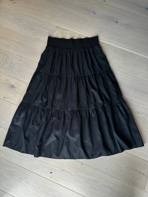 Kjol från Vintage Stories - Svart långkjol från Vintage Stories med bred, smockad midja och flera volanger. Kjolen är 100% polyester. Står tyvärr ingen storlek men gissningsvis är det storlek M/L.   Obs: Alla plagg säljs endast fram till 28/12 