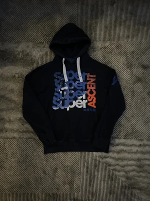 Mörkblå Superdry hoddie  - Svart Superdry hoodie med stor tryckt text i blått, vitt och rött på framsidan. Tröjan har huva med vita snören och en klassisk känguruficka. Märket Super Winter Sports Division syns i nacken. Perfekt för dig som gillar sportig och avslappnad stil.