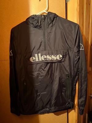 Svart vindjacka från Ellesse - Svart vindjacka från Ellesse med stor logga på bröstet, huva och half zip framtill. Jackan har två sidofickor med dragkedja och elastiska muddar vid ärmslut. Perfekt för dig som gillar sportig och clean stil. Är storlek S/36 men är väldigt liten i storlek, skulle säga att den mer är som XS! Säljes då den är för liten!